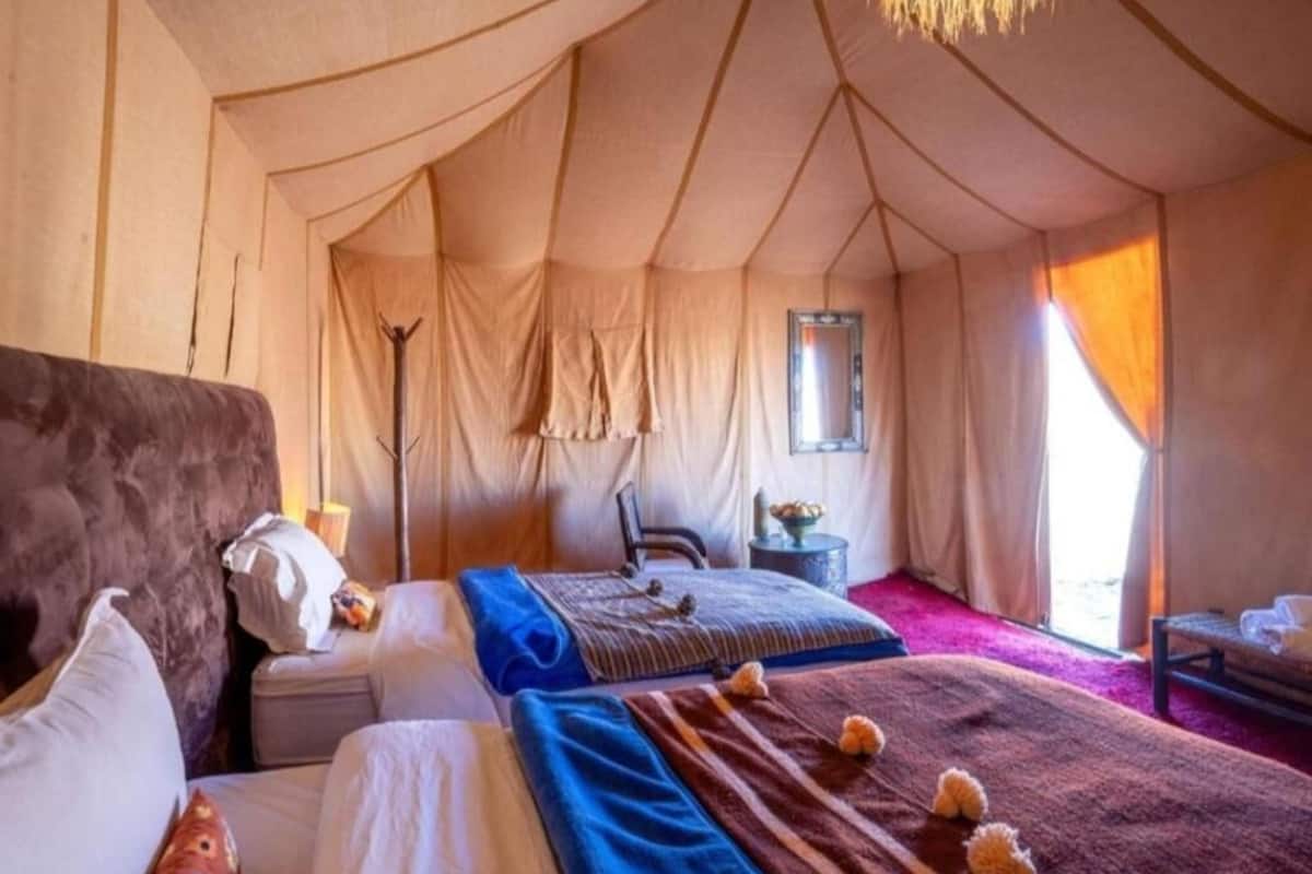 Quarto em tenda com duas camas, uma cadeira, uma mesinha e uma janela.
