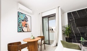 Un salon moderne avec une table ronde en bois, deux chaises, un coussin vert, un grand tableau abstrait et une porte-fenêtre coulissante donnant sur un balcon où se trouve un pot de fleurs.