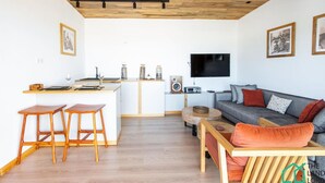 Una sala de estar moderna con techo de madera, un sofá gris con cojines naranja y una mesa de centro de madera, con un televisor montado en la pared.