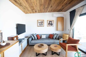 Un salon moderne avec un canapé gris, deux petites tables d’appoint rondes en bois, un téléviseur à écran plat fixé au mur et un plafond en bois.