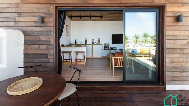 Un interior moderno con una pared de madera, una zona de comedor con una mesa redonda y sillas, una cocina con taburetes y una puerta corrediza de cristal que da a un espacio exterior con vistas a palmeras.