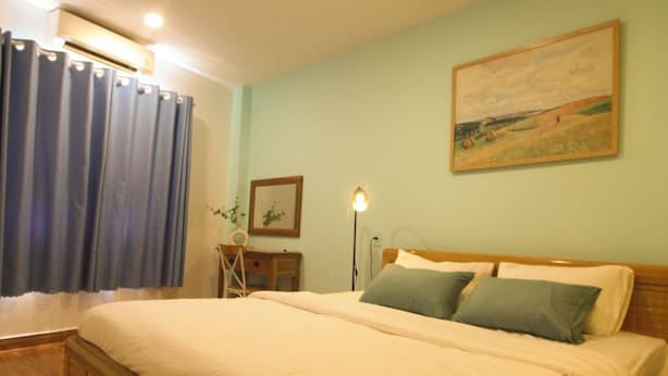 A bedroom with a wooden bed, a desk, a chair, a lamp, and a painting on the wall. - NHA NGHI AO (Ho Chi Minh City)