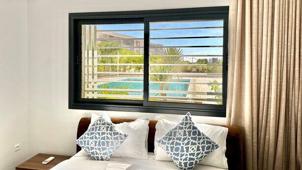 Ein Hotelzimmer mit einem Bett, zwei Kissen, einem Nachttisch, einem Fenster mit Blick auf einen Pool und Grünflächen, sowie einem Vorhang.