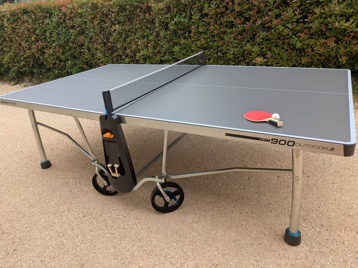 Una mesa de ping pong portátil con ruedas, identificada como 'PPT 900 Outdoor 2', ubicada al aire libre sobre una superficie de grava.