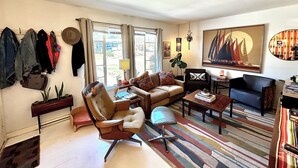 A living room with a sofa, armchair, and coffee table, a bookshelf, and a large window with curtains. - Mid century bungalow mins to Pearl, dt, airport (San Antonio)