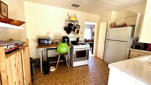 A compact kitchen with a microwave, stove, and refrigerator. A green chair is placed in front of the stove, and a small table with books is visible on the left. - Mid century bungalow mins to Pearl, dt, airport (San Antonio)