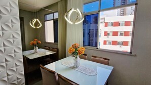 A modern hotel room with a dining table, chairs, a vase with flowers, a mirror, and a view of a building through the window. - COBAR0148 - Edifício Beta (Salvador)