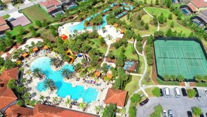 Vista aérea de un complejo turístico con una gran piscina, canchas de tenis y un aparcamiento.