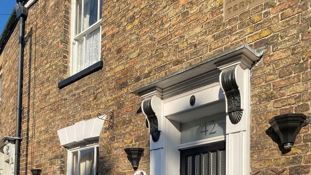 A brick building with a black door and white trim, a potted plant, and a small window with a curtain. - Serene 3-bedroom cottage in lovely Pocklington perfect for relaxing (Pocklington)