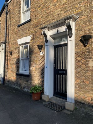 A brick building with a black door and white trim, a potted plant, and a small window with a curtain.