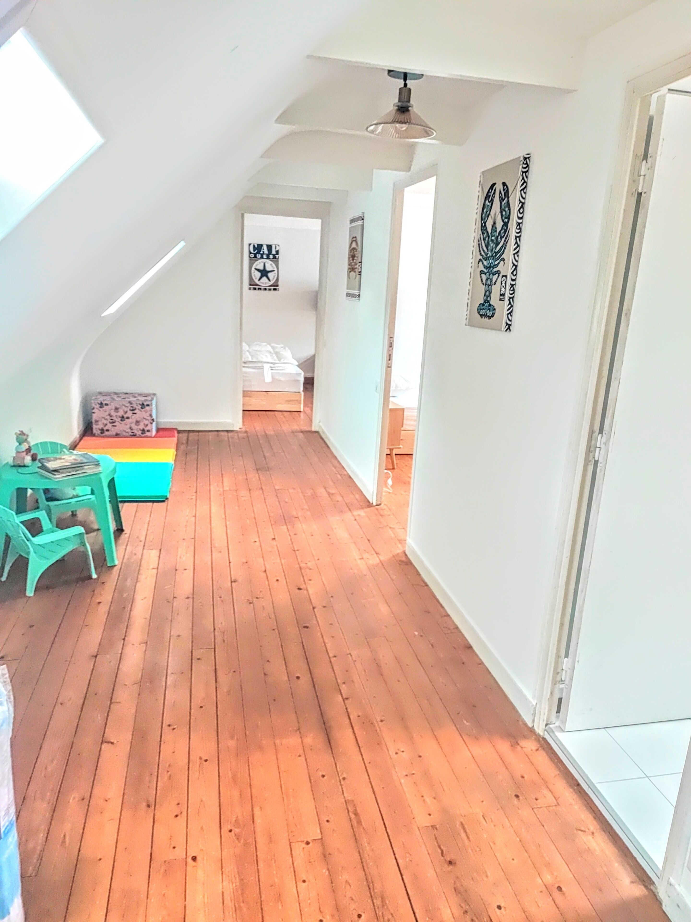 A narrow hallway with wooden flooring, a table with chairs, and a bed in the background.