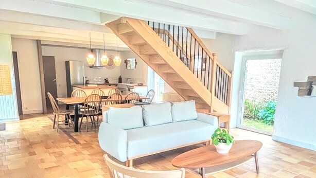 Amplia sala de estar con una escalera de madera, un sofá azul claro, una mesa de comedor con sillas y una pequeña mesa de madera con una planta.