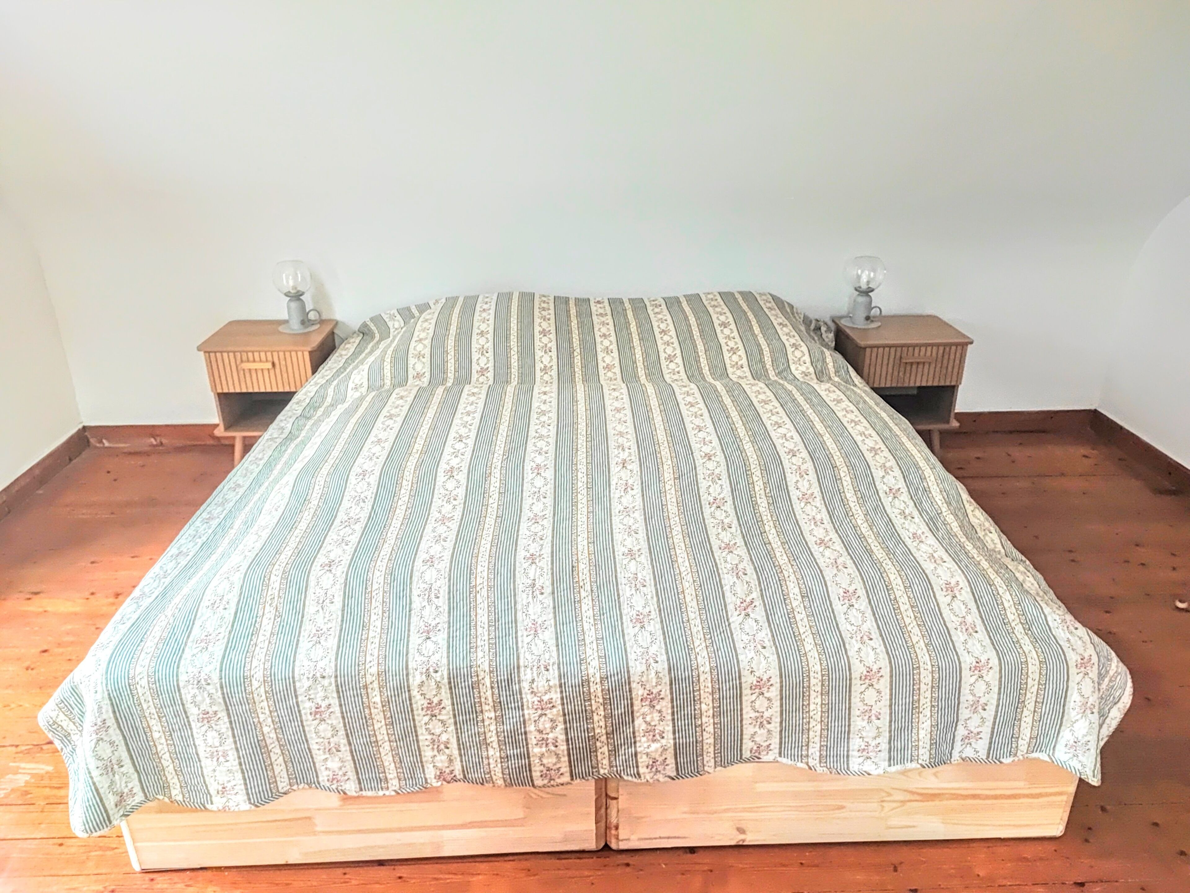 Una cama con una colcha a rayas y motivos florales, cabecera de madera y dos mesitas de noche con lámparas.