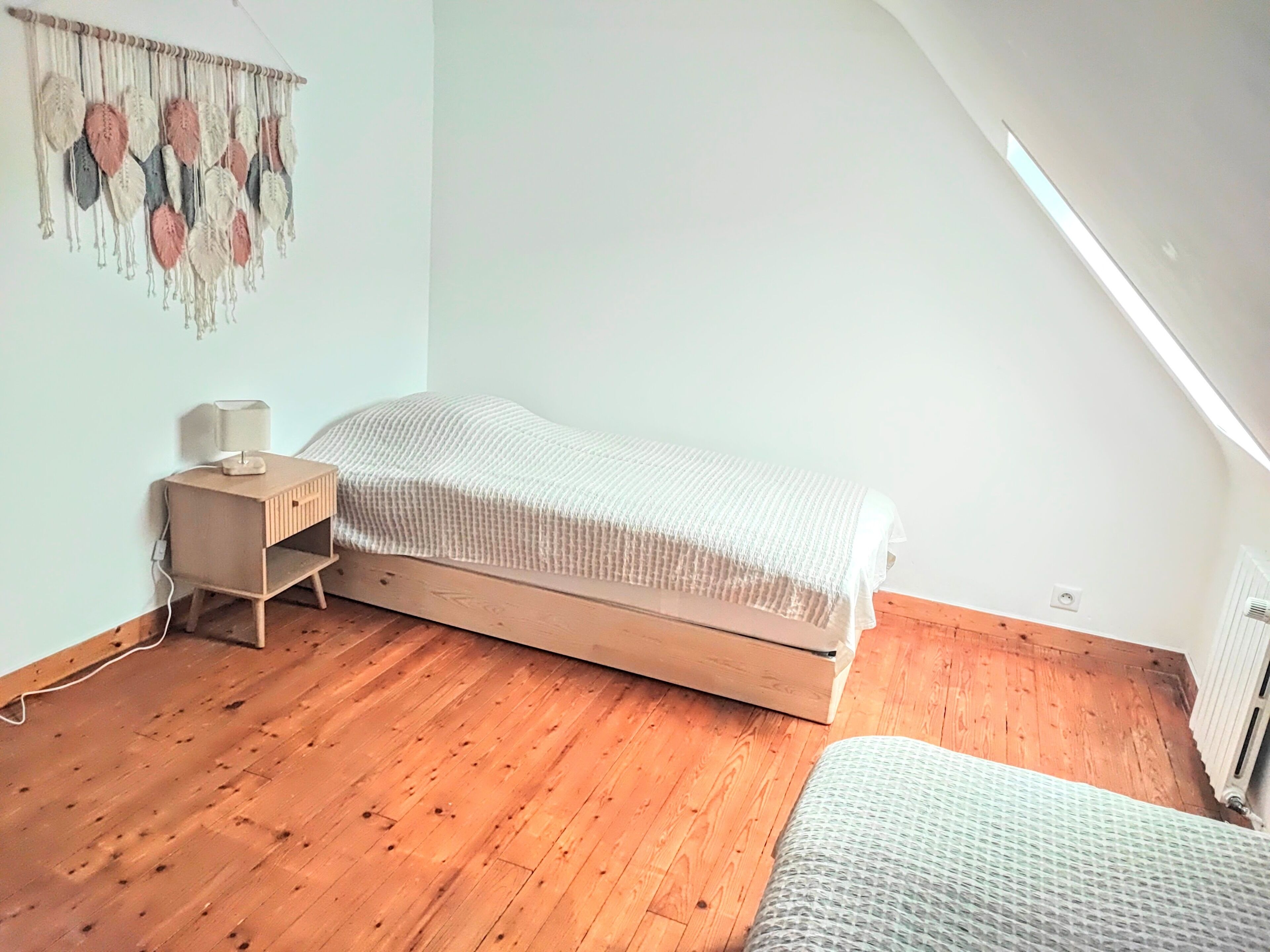 Une petite chambre à coucher, bien rangée, avec un plancher en bois, un lit simple, une table de chevet avec une lampe et une décoration murale.