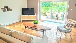Una sala de estar con un sofá, una mesa de centro con una planta, un televisor en un mueble bajo y una puerta corrediza de vidrio que da a un patio exterior con sillas y una mesa.