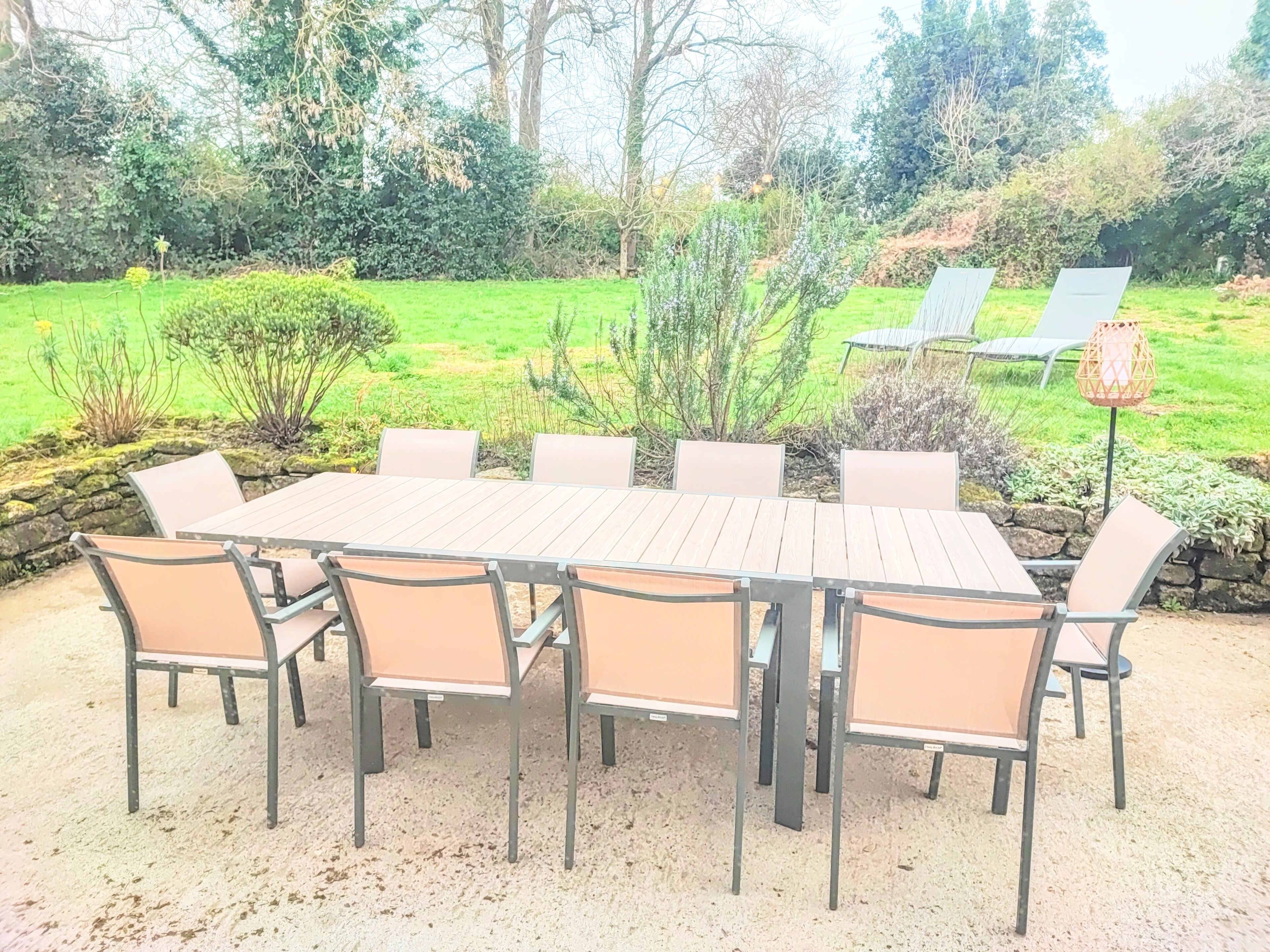 Ein rechteckiger Holztisch mit sechs Stühlen, umgeben von einer Steinmauer und einem Garten mit verschiedenen Pflanzen und Bäumen.