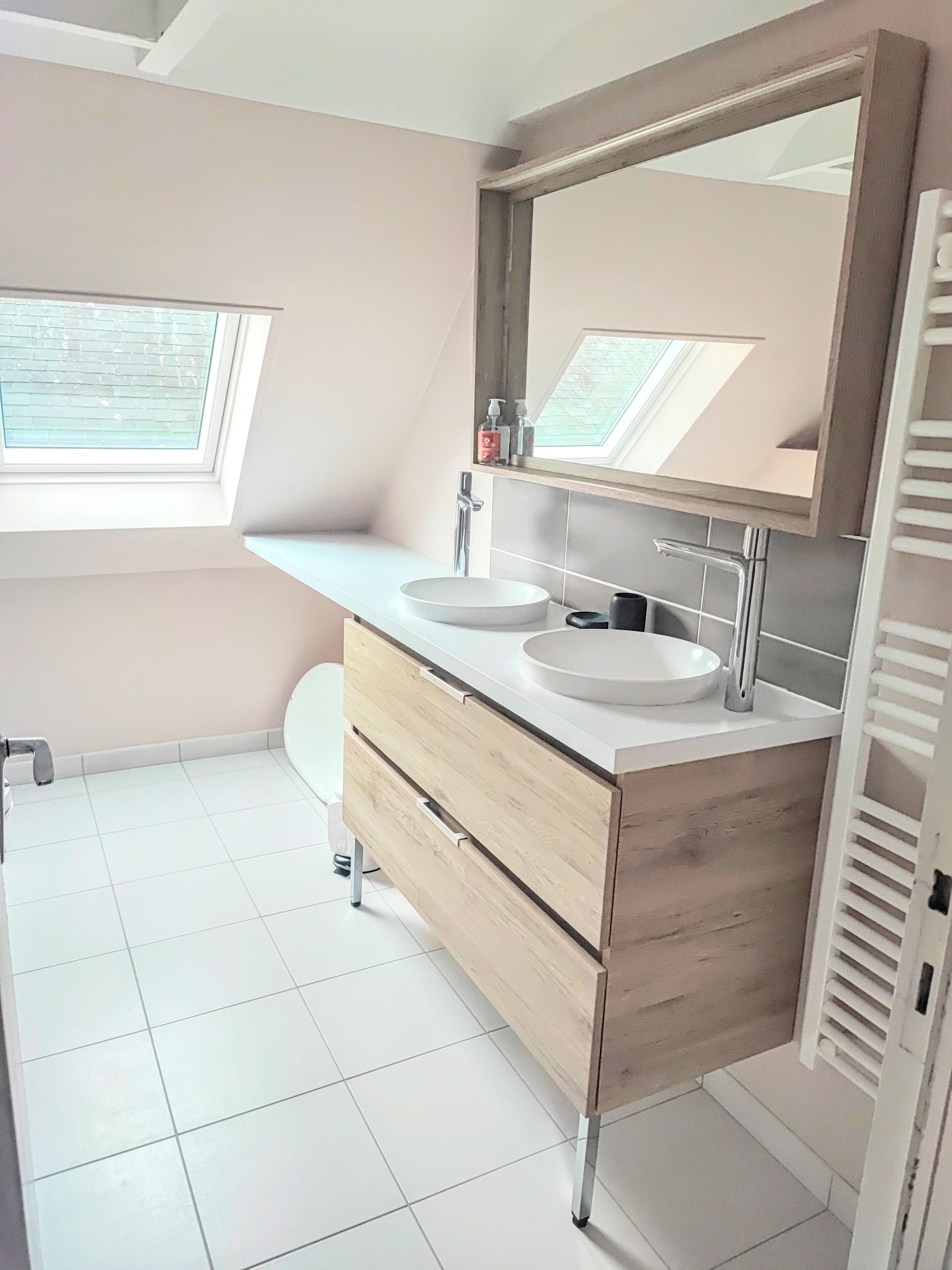A modern bathroom with a skylight, two sinks, a vanity with drawers, and a toilet.