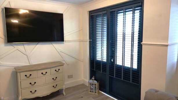 A room with a flat-screen TV mounted on the wall, a white dresser with multiple drawers, a black door with horizontal blinds, and a small white lantern on the floor. - Luxurious studio room in the heart of East Kilbride 10mins from a hospital (East Kilbride)