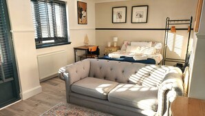 A cozy room with a grey sofa, a patterned rug, a bed with floral bedding, and a window with blinds. - Luxurious studio room in the heart of East Kilbride 10mins from a hospital (East Kilbride)