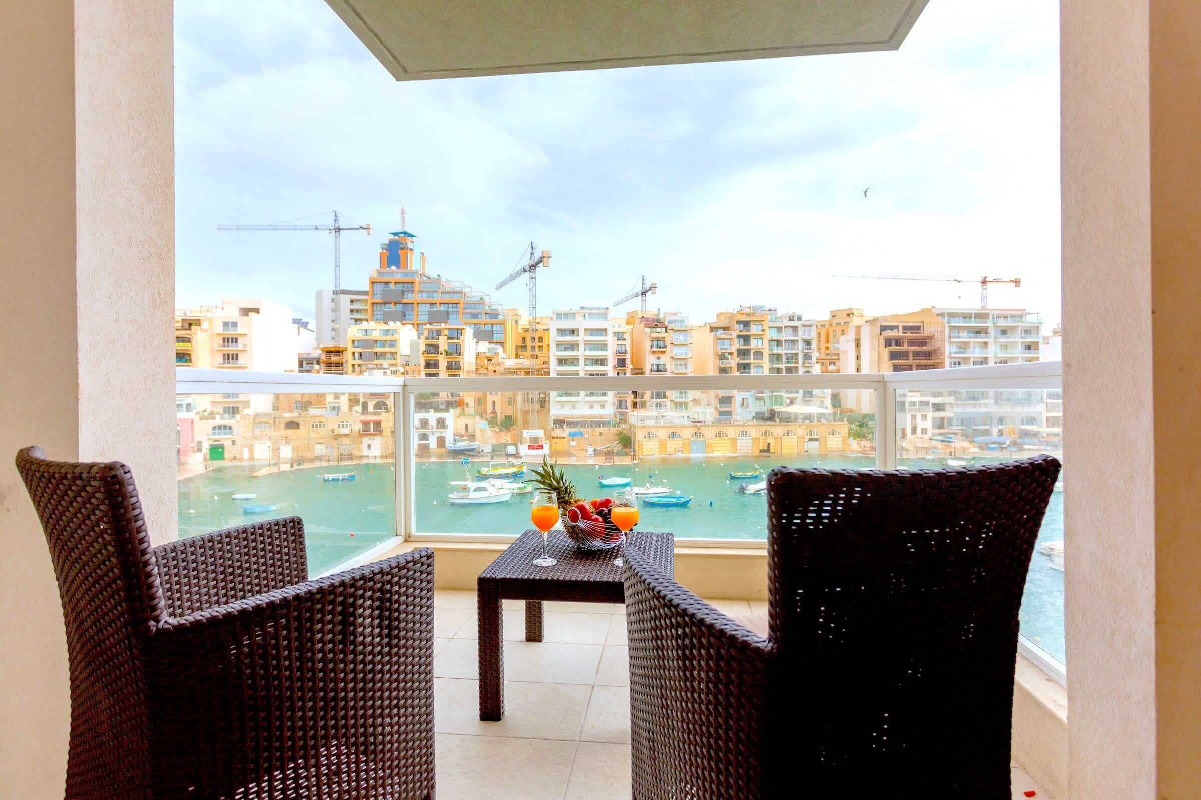 Een balkon met rieten stoelen en een tafel, met uitzicht op een jachthaven met boten en gebouwen op de achtergrond.