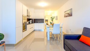 Une cuisine moderne avec des meubles blancs, un coin repas avec une table et des chaises blanches, et un canapé gris agrémenté d’un coussin orange.