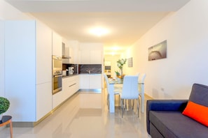 Une cuisine moderne avec des meubles blancs, un coin repas avec une table et des chaises blanches, et un canapé gris agrémenté d’un coussin orange.