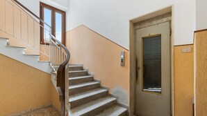 Uma escadaria com corrimão, uma porta com janela e um painel de comando de elevador fixado na parede.