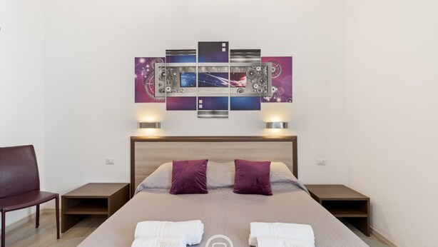 Quarto de hotel com cama, duas mesinhas de cabeceira, uma cadeira e uma obra de arte na parede.