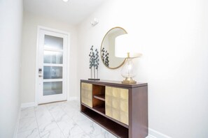 A hallway with a white door, a dark wooden console with glass doors, a round mirror, and a floor lamp. - Gorgeous 5 Bd Home Close to Disney   Storey Lake Resort 2734 (Kissimmee)