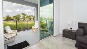 Uma sala de estar moderna com uma porta de vidro deslizante que dá para uma varanda com mobiliário para área externa e vista para um parque e edifícios.