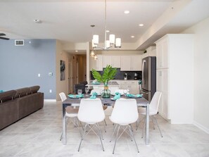 A modern dining area with a table set for four, white chairs, a chandelier, and a kitchen in the background. - Marvelous 2 Bd Apt Close to Disney at Storey Lake 106 3121 (Kissimmee)