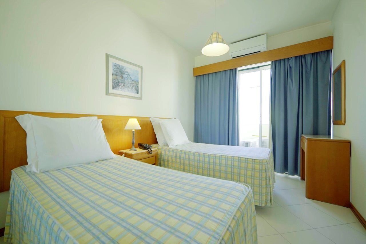 Camera d'albergo con due letti, un comodino con lampada, un tavolino e una finestra con tende.