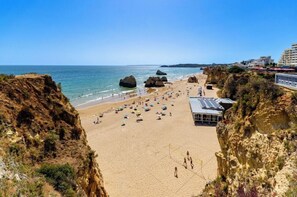 A beach with rocky cliffs, a sandy shore, and a building with a solar panel roof. - Turim Algarve Mor Apartamentos (Portimão)