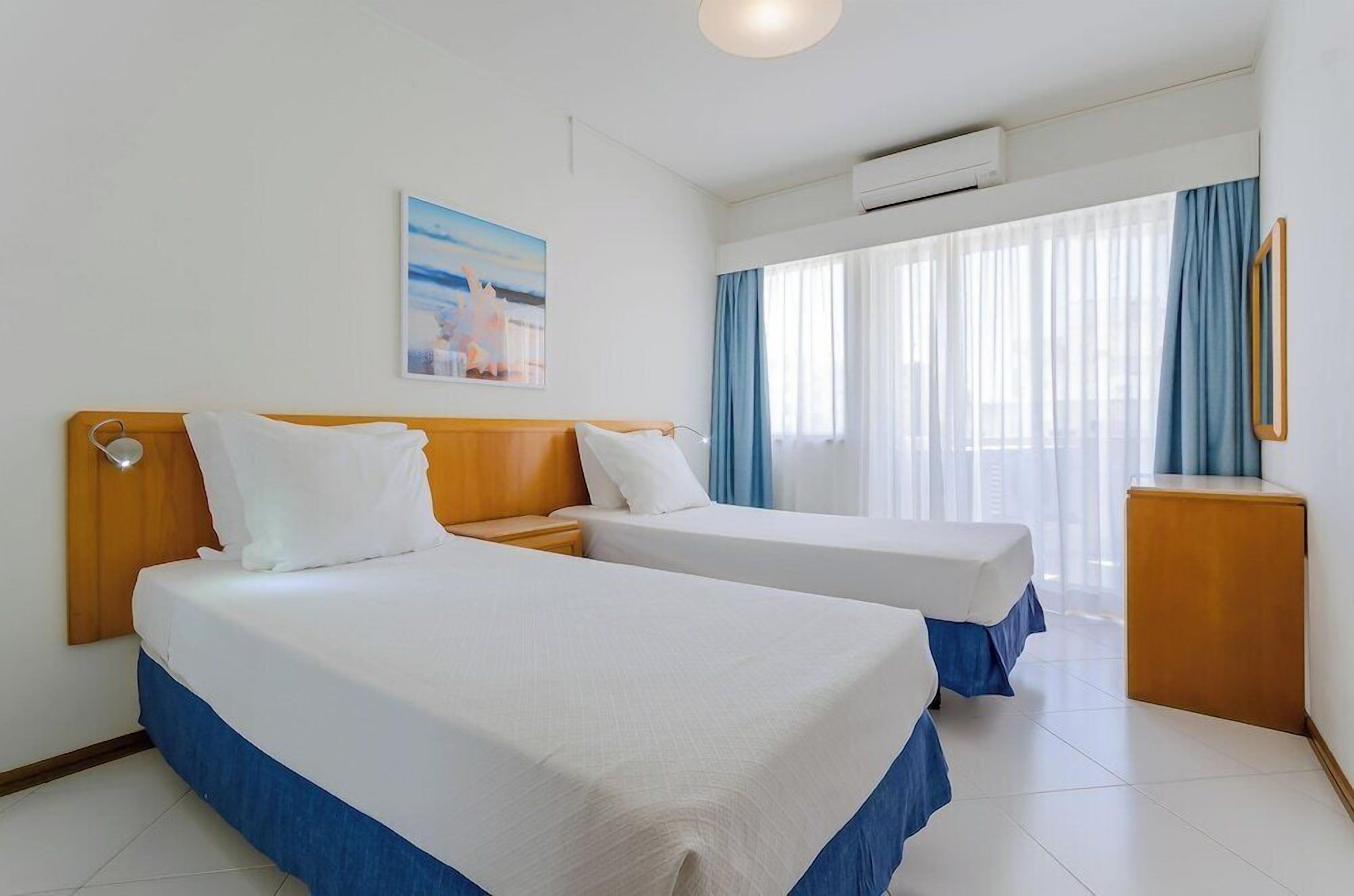 A hotel room with two single beds, a wooden headboard, a bedside table, a wall-mounted air conditioner, a window with sheer curtains, and a framed picture on the wall.