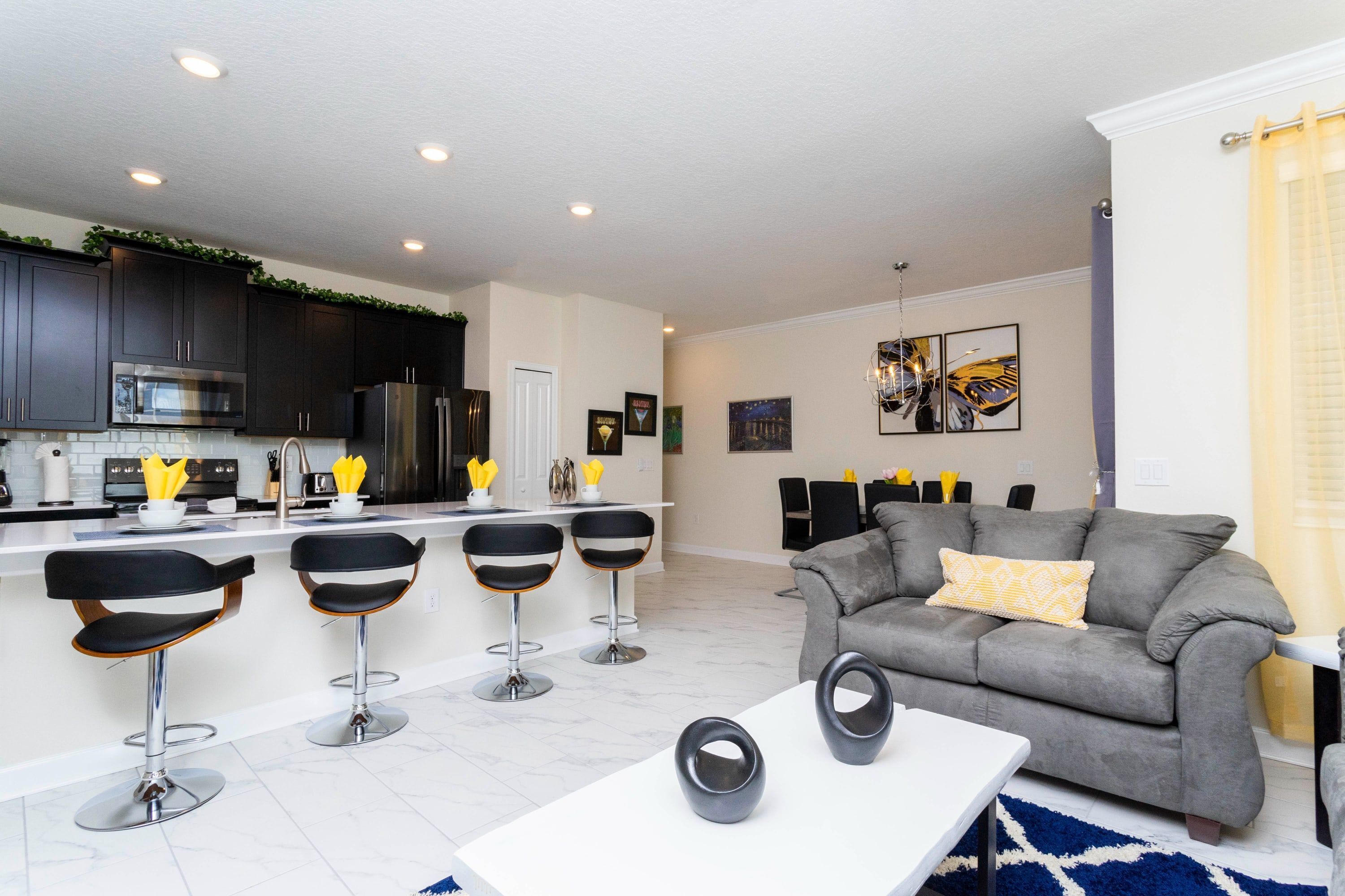 A modern kitchen with dark cabinets, a central island with bar stools, and a grey sofa in the living area.