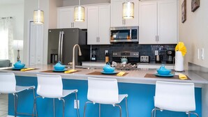 A modern kitchen with white cabinets, a stainless steel refrigerator, a microwave, and a sink. There are three white bar stools and a blue accent wall. - Stunning 5 Bd Home w  Pool Close to Disney 4822 (Kissimmee)