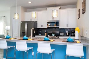 Een moderne keuken met witte kasten, een roestvrijstalen koelkast, een magnetron en een spoelbak. Drie witte barkrukken en een blauwe accentmuur.