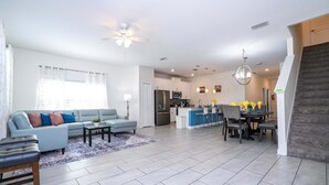 A spacious living area with a sofa, coffee table, and a dining table with chairs. A kitchen is visible in the background with a refrigerator, microwave, and sink. - Stunning 5 Bd Home w  Pool Close to Disney 4822 (Kissimmee)