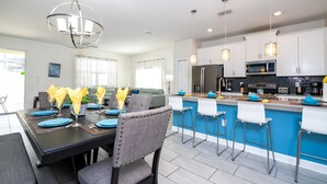 A modern kitchen with white cabinets, a blue island, and a dining area with a black table and grey chairs. - Stunning 5 Bd Home w  Pool Close to Disney 4822 (Kissimmee)
