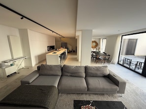 A modern living room with a grey sofa, a small coffee table, and a keyboard on a stand. There is a kitchen area with a dining table and chairs visible in the background. - Large modern house in the center of town (Paris)