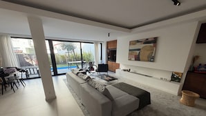 A modern living room with a large grey sectional sofa, a coffee table, and a dining area with a table and chairs. There is a large window providing a view of the outdoors. - Large modern house in the center of town (Paris)
