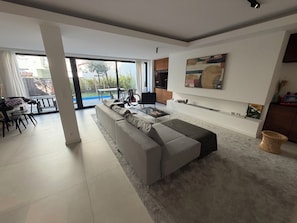 A modern living room with a large grey sectional sofa, a coffee table, and a dining area with a table and chairs. There is a large window providing a view of the outdoors. - Large modern house in the center of town (Paris)