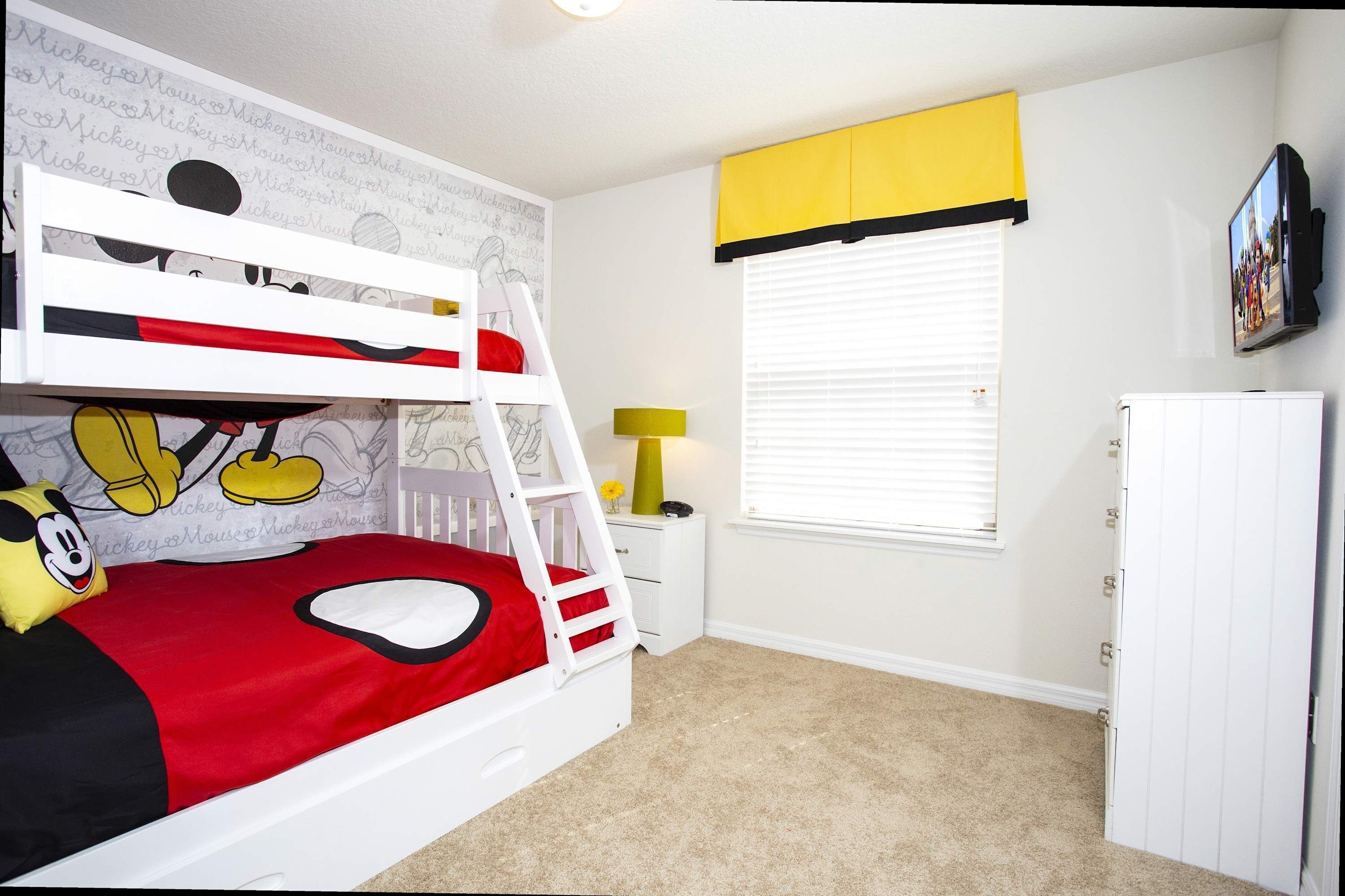 A bunk bed with Mickey Mouse-themed bedding, a small bedside table with a lamp, a window with blinds, and a TV mounted on the wall.