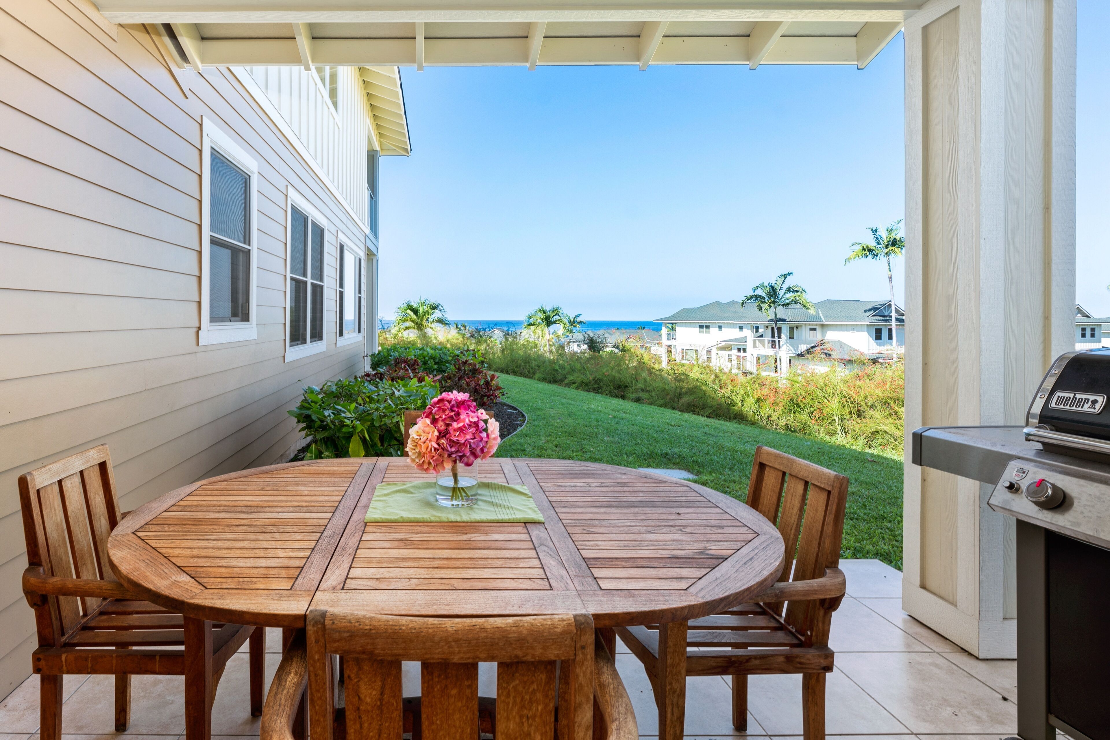 A wooden patio table with chairs, a grill, and a view of the ocean and houses in the background.