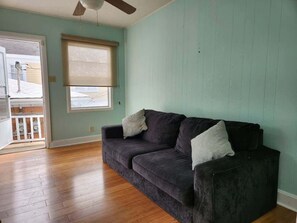 Ein Zimmer mit einem dunkelbraunen Sofa, zwei Kissen, einem Fenster mit einer beigen Jalousie und Holzboden.