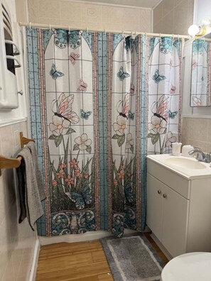 A bathroom with a shower curtain featuring floral and butterfly designs, a wooden floor, a sink, and a mirror. - Cozy Church Street Apartment In Historic Galveston 2 Bedroom Apts (Galveston)