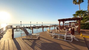 A wooden deck with lounge chairs and a covered seating area overlooking a calm body of water. - Direct Bayfront Sunsets - Boat Docks - Pool - Fish 2 Bedroom Apts (South Padre Island)