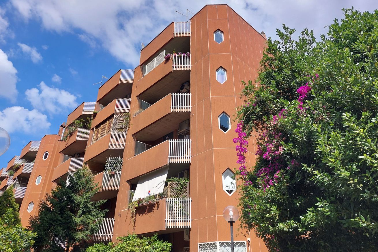 Um edifício residencial com vários andares, varandas, céu claro e vegetação ao redor.
