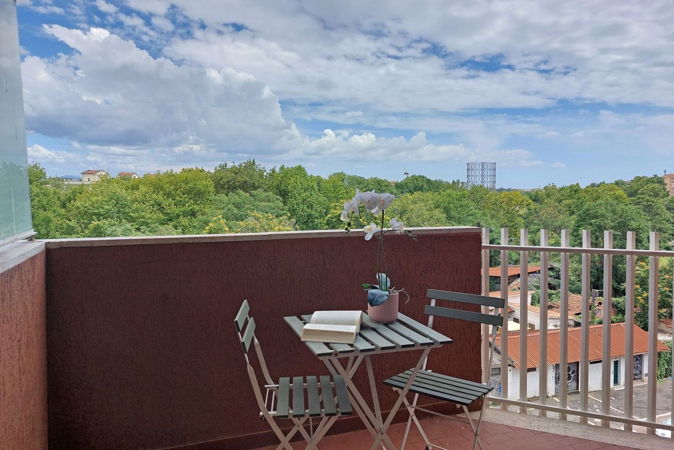 Ein Balkon mit Tisch und Stühlen, einem Blumentopf und Blick auf eine grüne Landschaft und Gebäude.