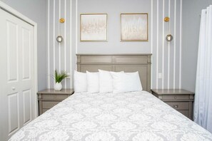 Un dormitorio ordenado con una cama grande, dos mesitas de noche, una lámpara de noche, una planta en maceta y dos cuadros en la pared.